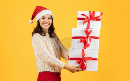 Foto de Mujer alegre con sombrero rojo de Papá Noel sosteniendo una pila de regalos de Año Nuevo y Navidad, sonriendo de alegría, posando en un estudio amarillo, epítome de la temporada festiva y dando espíritu. Oferta de Navidad ad - Imagen libre de derechos