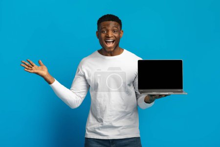 Téléchargez les photos : Jeune homme noir excité montrant ordinateur portable avec écran blanc isolé sur fond bleu. Publicité, grande offre, site Web et recommandation de professionnels. Travail à distance, freelance - en image libre de droit