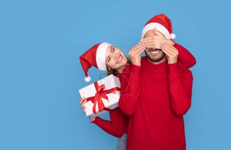Téléchargez les photos : Joyeux Noël. Jeune femme surprenant son petit ami avec un cadeau de Noël couvrant ses yeux et souriant à la caméra, couple portant des chapeaux de Père Noël célébrant les vacances ensemble, posant sur fond bleu - en image libre de droit