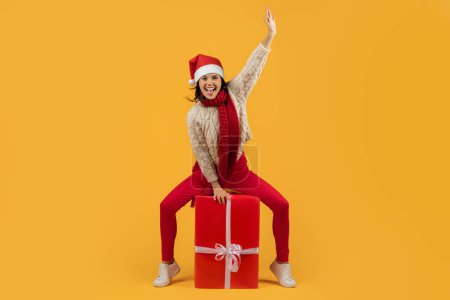 Téléchargez les photos : Joyeux jeune femme dans le chapeau du Père Noël assis sur un grand cadeau de Noël enveloppé, pour les achats de vacances et les annonces de célébration, studio tourné sur fond jaune. Longueur totale - en image libre de droit