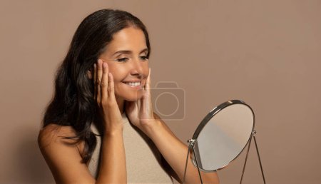 Foto de Alegre señora madura bastante europea con maquillaje natural, disfrutar del cuidado de la piel, mirar el espejo, aislado en el fondo del estudio beige, panorama. Cuidado de la belleza, cosméticos, anuncios y ofertas - Imagen libre de derechos