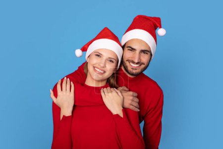 Photo for Cheerful European Couple Wearing Red Sweaters And Santa Hats Posing On Blue Background, Happy Romantic Spouses Embracing And Smiling At Camera, Having Fun Together, Enjoying Christmas Holidays - Royalty Free Image