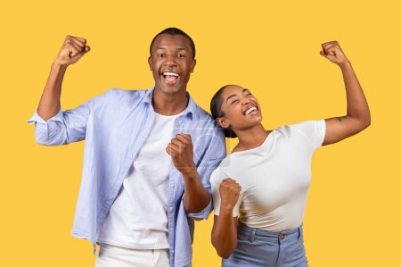 Téléchargez les photos : Homme et femme noirs joyeux avec des poings levés célébrant le succès, les yeux fermés avec de larges sourires, portant des vêtements décontractés sur fond jaune - en image libre de droit