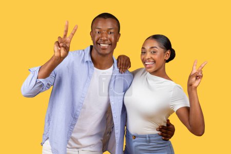 Téléchargez les photos : Jeune couple noir rayonnant souriant largement, faisant des signes de paix avec leurs mains sur fond jaune monochrome, souriant à la caméra - en image libre de droit