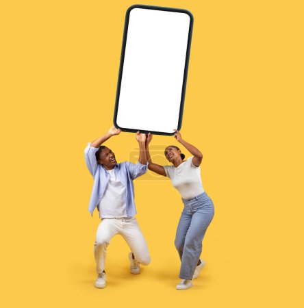 Téléchargez les photos : Jeunes conjoints noirs enthousiastes tiennent avec confiance téléphone cellulaire massif avec écran blanc, créant une humeur promotionnelle pour une offre passionnante, sur fond jaune vif - en image libre de droit