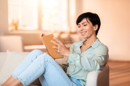 Téléchargez les photos : Femme brune souriante absorbée par la lecture de fiction, confortablement assise sur son canapé avec un livre en papier, incarnant une pause paisible le week-end, entourée par le confort de son intérieur - en image libre de droit