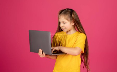 Téléchargez les photos : Portrait de curieuse préadolescente utilisant un ordinateur portable tout en se tenant isolé sur fond rose, heureuse enfant femelle regardant l'écran d'ordinateur avec intérêt, navigation sur le site éducatif, espace de copie - en image libre de droit
