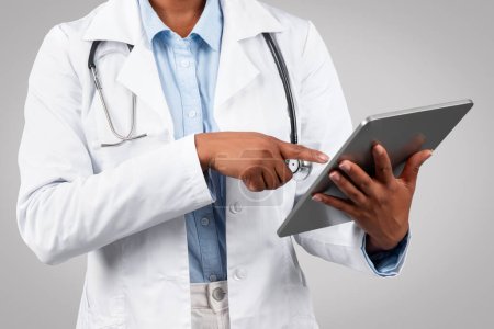 Foto de Milenial terapeuta médica mujer negro en bata blanca, charlando en la tableta, escribiendo en gadget, aislado en el estudio de fondo gris, recortado. Aplicación de atención médica, medicina moderna - Imagen libre de derechos