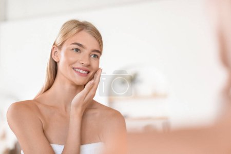 Téléchargez les photos : Heureuse jeune femme aux cheveux blonds touchant sa peau douce avec soin, posant enveloppée dans une serviette blanche, se livre à des soins du visage choyants dans sa salle de bain moderne à la maison - en image libre de droit