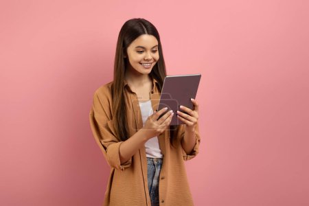 Téléchargez les photos : Souriant adolescent fille regardant l'écran de tablette numérique avec intérêt, heureuse adolescente appréciant le contenu en ligne tout en utilisant un gadget moderne, debout sur fond de studio rose, espace de copie - en image libre de droit