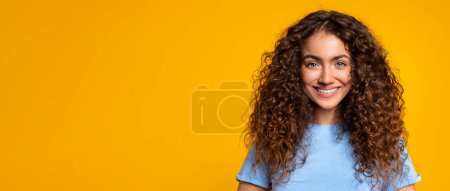 Photo for Radiant young woman with lush curly hair smiling warmly, wearing blue t-shirt against vivid yellow background, exuding positivity and confidence in headshot portrait, panorama, copy space - Royalty Free Image