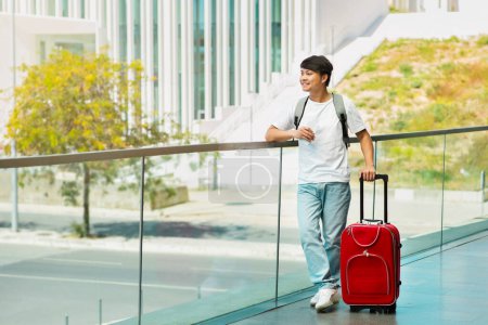 Photo for Cheerful young asian man in casual outfit tourist with suitcase and phone in his hand standing outdoors next to airport or train station, waiting for taxi, looking at copy space - Royalty Free Image