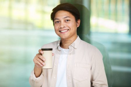 Téléchargez les photos : Portrait d'homme asiatique millénaire joyeux et joyeux portant une tenue décontractée buvant du café à emporter le matin au centre d'affaires du centre-ville sur le chemin du bureau, souriant à la caméra - en image libre de droit