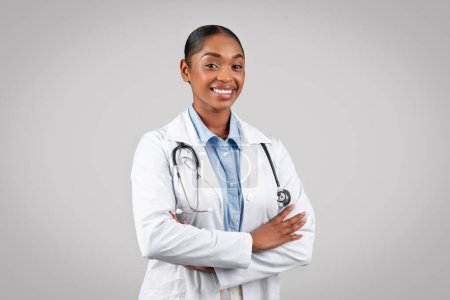 Photo for Professional cheerful black doctor smiling at camera, with arms crossed, in white coat and stethoscope on grey background, studio. Medical health care, work and therapist help - Royalty Free Image