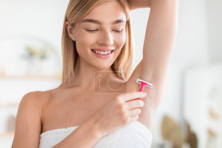 Téléchargez les photos : Jeune femme blonde se rase les aisselles à l'aide d'un rasoir dans la salle de bain, en se concentrant sur l'hygiène de l'épilation du corps et une peau lisse dans sa routine quotidienne, debout avec le bras levé - en image libre de droit