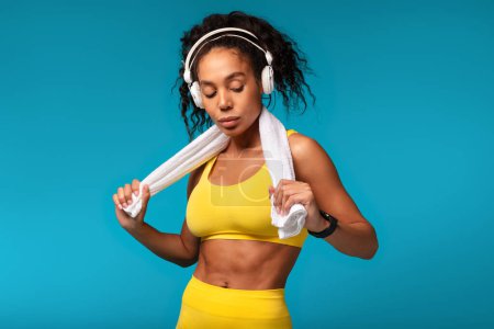 Photo for Workout Motivation. Determined Black Fitness Lady Wearing Wireless Headphones Standing With Towel After Training Over Blue Background. Studio Shot Of Athletic Woman In Yellow Fitwear - Royalty Free Image