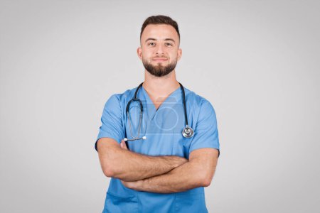 Téléchargez les photos : Jeune médecin européen vêtu d'un manteau bleu, les bras croisés, respirant confiance et professionnalisme, capturé sur fond gris minimaliste - en image libre de droit
