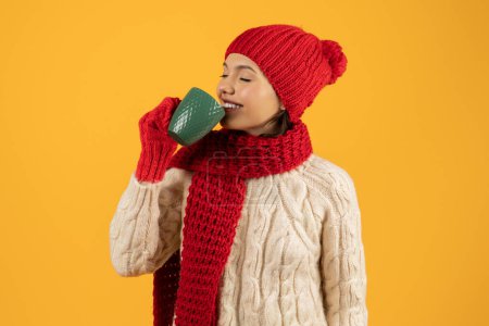 Photo for Joyful lady in knitted hat and gloves, savoring a warm drink, eyes closed in pleasure, modern holiday concept, studio shot on yellow backdrop with space for New Year offers - Royalty Free Image
