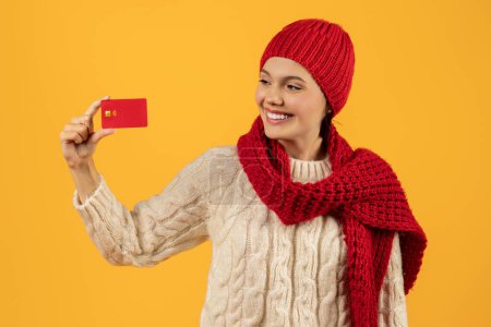 Photo for Smiling young lady in red knitwear and scarf holding credit card, ready for holiday shopping, perfect for finance and sale ads, studio shot against yellow background. Bank winter offers - Royalty Free Image