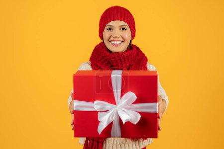 Foto de Oferta de Navidad. Mujer alegre en traje de invierno que muestra el regalo de Año Nuevo envuelto, la celebración de la caja de regalo roja, perfecto para los anuncios de compras de vacaciones, estudio de tiro contra el fondo amarillo - Imagen libre de derechos