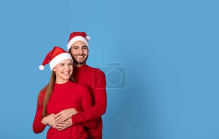 Téléchargez les photos : Joyeux jeune couple dans Santa chapeaux étreignant sur fond de studio bleu, bel homme embrassant sa belle petite amie par derrière, conjoints aimants célébrant le Nouvel An ou Noël ensemble, espace de copie - en image libre de droit