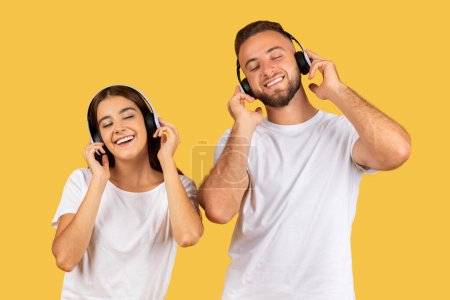 Photo for Cheerful millennial european couple in white t-shirts, wireless headphones, listen music, enjoy song, rest, isolated on yellow studio background. Free time, online audio app, relax - Royalty Free Image