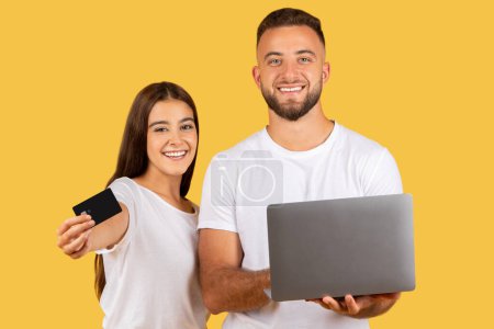 Photo for Happy millennian european wife and husband in white t-shirts with computer show credit card, isolated on yellow studio background. Ad and offer, profit banking, sale recommendation, online shopping - Royalty Free Image