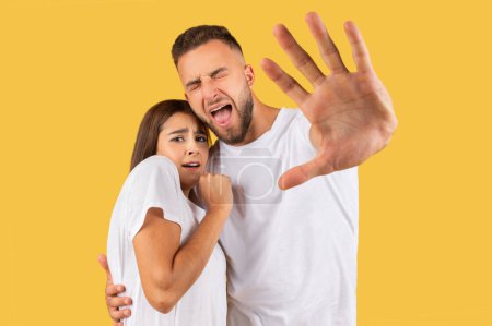Téléchargez les photos : Peur jeune couple européen triste en t-shirts blancs faire geste d'arrêt à la main, isolé sur fond de studio jaune. Effrayé et terrifié, expression de la peur, choc et panique - en image libre de droit