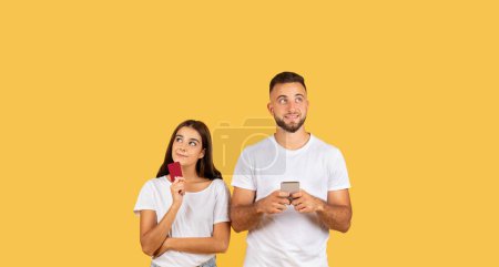 Photo for Positive pensive young european family in white t-shirts, with credit card, phone think, dream about purchases, isolated on yellow studio background, panorama. Add and offer, online sale, shopping - Royalty Free Image
