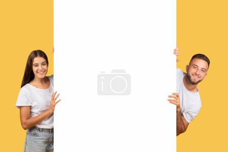 Photo for Happy millennial european couple in white t-shirts with big banner with empty space, isolated on yellow studio background. Ad and offer attention, sale, ad and offer, good news - Royalty Free Image