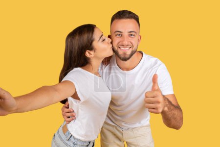 Photo for Positive young caucasian lady in white t-shirt kiss man in check, have fun, enjoy date, show thumb up sign, make selfie on phone, isolated on yellow studio background. Love approve, photo for blog - Royalty Free Image