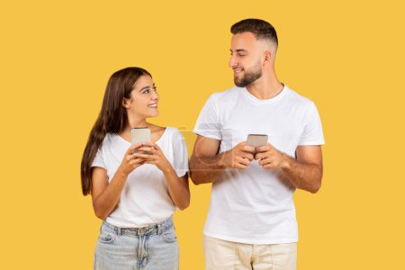 Photo for Smiling millennial european lady and guy in white t-shirts typing on phone, chatting at gadget, isolated on yellow studio background, close up. Shopping, communication online app - Royalty Free Image