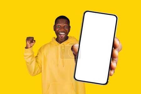 Téléchargez les photos : Homme noir dans la célébration avec les poings tremblants, tenant grand téléphone avec écran vide pour la maquette, symbolisant la joie et la victoire, parfait pour la technologie et les concepts de succès, fond studio jaune - en image libre de droit