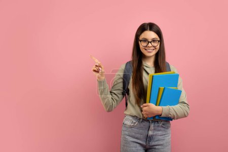 Photo for Happy teen girl holding books and pointing aside at copy space, female student wearing glasses and backpack indicating free place for educational advertisement, standing against pink background - Royalty Free Image