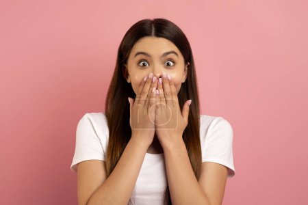 Photo for Surprised teen girl covering mouth with hands and looking at camera, shocked teenage female standing with wide open eyes, expressing astonishment, posing against pink background, free space - Royalty Free Image