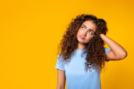 Photo for Pensive young woman with curly hair looking up at free space, hand in hair, lost in thought against solid yellow background, symbolizing curiosity or contemplation - Royalty Free Image