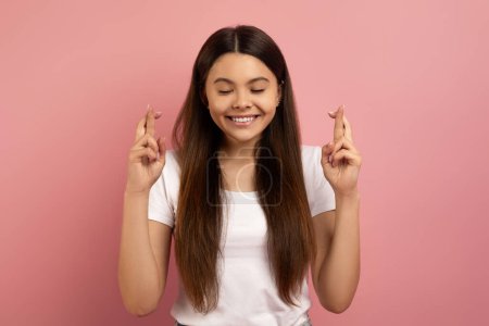 Photo for Hopeful teen girl crossing fingers and making wish, superstitious smiling female teenager pleading for good luck and fortune, standing with eyes closed against pink studio background, copy space - Royalty Free Image