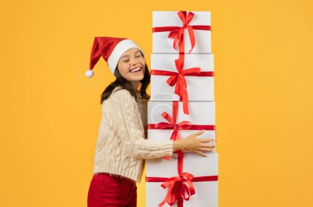 Téléchargez les photos : Joyeux jeune femme dans le chapeau Santa embrassant pile de cadeaux de Noël avec joie, studio tourné sur fond jaune vif. Dame avec de nombreux cadeaux pour les offres de la saison des fêtes - en image libre de droit
