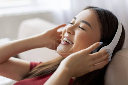 Photo for Happy Young Asian Woman In Wireless Headphones Listening Music At Home, Closeup Shot Of Cheerful Korean Lady Relaxing On Couch With Closed Eyes, Enjoying Favorite Playlist, Side View - Royalty Free Image