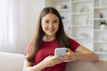 Photo for Beautiful Smiling Asian Woman With Smartphone In Hands Sitting On Couch At Home, Happy Young Korean Female Using Mobile Phone For Communication Or Online Shopping, Looking At Camera, Copy Space - Royalty Free Image