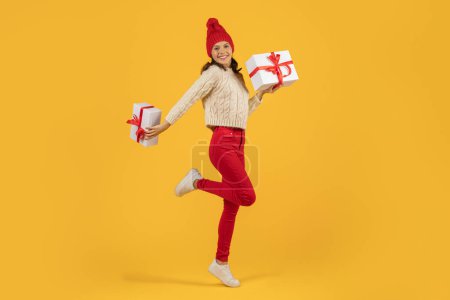 Téléchargez les photos : Joyeuse femme en tricot rouge sautant chapeau avec Noël cadeaux de vacances, posant plein de joie, studio tourné sur fond jaune, idéal pour Noël et Nouvel An offres - en image libre de droit