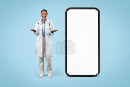 Téléchargez les photos : Joyeux médecin noir jeune femme en manteau blanc, tenir l'espace libre sur les mains près du grand téléphone avec écran vide, isolé sur fond bleu studio. Soins médicaux choisir - en image libre de droit