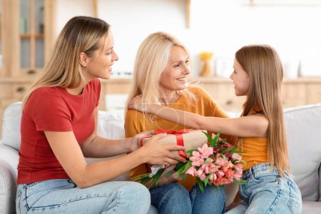 Photo for Loving grown up daughter and preteen granddaughter kissing and hugging, greeting granny with International Womens Day, excited mature lady holding present, flowers, home interior - Royalty Free Image