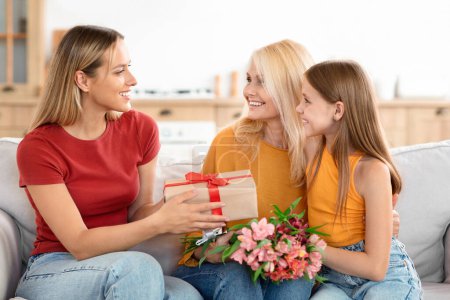 Photo for Three generations of women on Mothers Day at home. A cheerful family moment as a grandmother is greeted with International Womens Day gifts by her daughter and granddaughter - Royalty Free Image