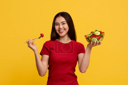 Photo for Healthy food, nutrition, diet. Positive pretty young taiwanese woman wearing red t-shirt holding fork and bowl with fresh vegetable salad, smiling at camera isolated on yellow studio background - Royalty Free Image