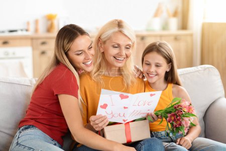 Photo for Adoring adult daughter and her young granddaughter embrace their grandmother, joyously celebrating Mothers Day, with the elderly woman receiving gifts, a bouquet, and a homemade card. - Royalty Free Image