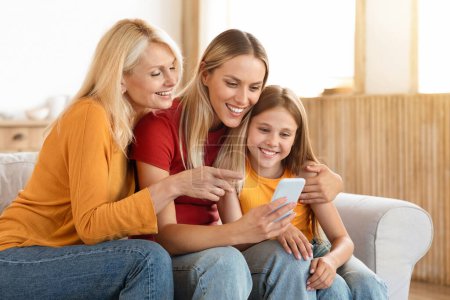 Téléchargez les photos : Mère, fille et petite-fille utilisent le téléphone ensemble à la maison. Trois générations de femmes partagent un rire sur un smartphone, capturant un moment de famille joyeux et connecté, faisant du shopping en ligne - en image libre de droit