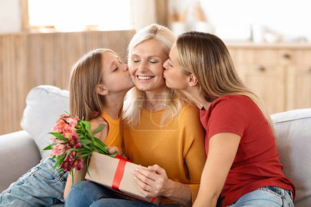 Photo for Adoring adult daughter and her young granddaughter kissing their grandmother, joyously celebrating International Womens Day, with the elderly woman receiving gift and bouquet, home interior - Royalty Free Image