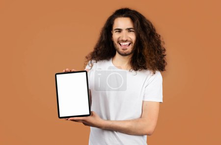 Téléchargez les photos : Jeune homme hispanique aux cheveux longs bouclés démontre tablette numérique avec écran d'ordinateur vide, mettant en valeur le site e-learning pour l'enseignement à distance, debout sur fond beige. Maquette - en image libre de droit
