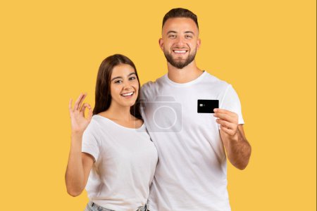 Photo for Positive young european family in white t-shirts, with credit card hugs, show ok sign with hand, isolated on yellow studio background. Recommendation shopping, cashback, profit - Royalty Free Image